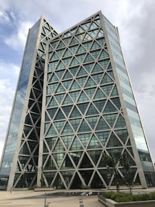 A modern architectural building with a distinctive crisscross geometric pattern, consisting of two tall, mirrored towers joined by a central structure. The facade features glass panels, and the surrounding area is landscaped with small trees and paths under a cloudy sky.
