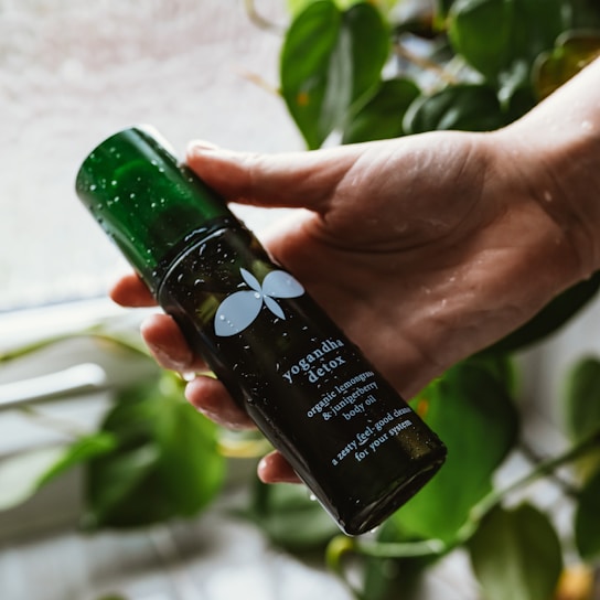 A hand is holding a dark green bottle labeled 'yogandha detox,' which contains organic lemongrass and juniper berry body oil. The bottle is wet, suggesting a fresh or natural setting, and in the background, there are green leafy plants, providing an organic and calming atmosphere.