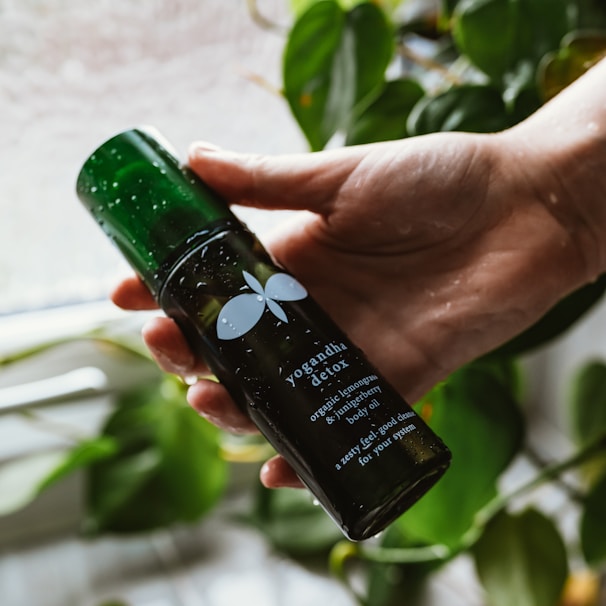 A hand is holding a dark green bottle labeled 'yogandha detox,' which contains organic lemongrass and juniper berry body oil. The bottle is wet, suggesting a fresh or natural setting, and in the background, there are green leafy plants, providing an organic and calming atmosphere.