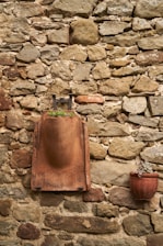 A rustic stone wall with a mix of irregularly shaped rocks. A terracotta-colored sculpture is mounted on the wall, featuring a rounded, abstract form with a small plant growing at its base. Adjacent to it, a red clay pot containing a small succulent hangs slightly lower on the wall.
