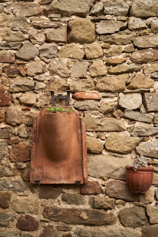 A rustic stone wall with a mix of irregularly shaped rocks. A terracotta-colored sculpture is mounted on the wall, featuring a rounded, abstract form with a small plant growing at its base. Adjacent to it, a red clay pot containing a small succulent hangs slightly lower on the wall.