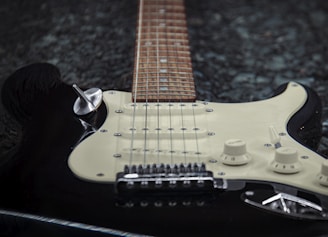 An electric guitar lays on a textured dark surface, featuring a cream-colored pickguard, six strings, and control knobs. The viewpoint is from the body's angle, highlighting the wooden fretboard leading off into the distance.