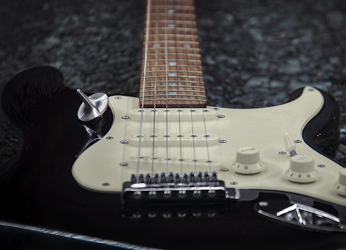 An electric guitar lays on a textured dark surface, featuring a cream-colored pickguard, six strings, and control knobs. The viewpoint is from the body's angle, highlighting the wooden fretboard leading off into the distance.