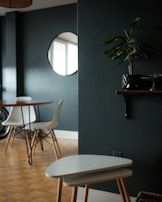 A modern interior room with a minimalistic design. There is a round wooden table with white chairs around it, set against a dark green wall. A round mirror is mounted on the wall, reflecting part of the window. A small potted plant sits on a wall shelf, and a pair of nesting tables are positioned in the foreground on a wooden parquet floor.