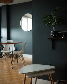 A modern interior room with a minimalistic design. There is a round wooden table with white chairs around it, set against a dark green wall. A round mirror is mounted on the wall, reflecting part of the window. A small potted plant sits on a wall shelf, and a pair of nesting tables are positioned in the foreground on a wooden parquet floor.