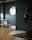 A modern interior room with a minimalistic design. There is a round wooden table with white chairs around it, set against a dark green wall. A round mirror is mounted on the wall, reflecting part of the window. A small potted plant sits on a wall shelf, and a pair of nesting tables are positioned in the foreground on a wooden parquet floor.