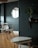 A modern interior room with a minimalistic design. There is a round wooden table with white chairs around it, set against a dark green wall. A round mirror is mounted on the wall, reflecting part of the window. A small potted plant sits on a wall shelf, and a pair of nesting tables are positioned in the foreground on a wooden parquet floor.