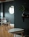 A modern interior room with a minimalistic design. There is a round wooden table with white chairs around it, set against a dark green wall. A round mirror is mounted on the wall, reflecting part of the window. A small potted plant sits on a wall shelf, and a pair of nesting tables are positioned in the foreground on a wooden parquet floor.
