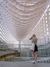 A person stands in an architectural setting with a complex, curved ceiling structure made of glass and steel. They are holding a camera, looking towards the roof. The space is filled with natural light, highlighting the modern and intricate design of the interior.