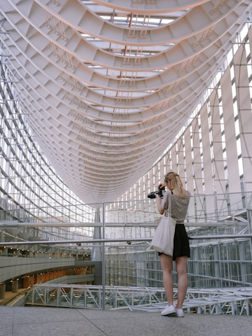 A person stands in an architectural setting with a complex, curved ceiling structure made of glass and steel. They are holding a camera, looking towards the roof. The space is filled with natural light, highlighting the modern and intricate design of the interior.