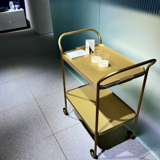 A gold-colored metal cart with two shelves is positioned against a textured glass wall. The top shelf holds two transparent cups and some papers, likely brochures or menu cards. The surface beneath the cart consists of large gray tiles with a slightly speckled texture. In the background, part of a modern, minimalist interior is visible, featuring additional shelves or storage units.