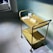 A gold-colored metal cart with two shelves is positioned against a textured glass wall. The top shelf holds two transparent cups and some papers, likely brochures or menu cards. The surface beneath the cart consists of large gray tiles with a slightly speckled texture. In the background, part of a modern, minimalist interior is visible, featuring additional shelves or storage units.