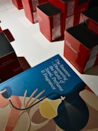 A collection of red and black boxes are neatly arranged on a white surface. In the foreground, there's a blue book titled 'The Essence Discovering the World of Scent, Perfume & Fragrance' with abstract art featuring a stylized perfume bottle and decorative motifs.