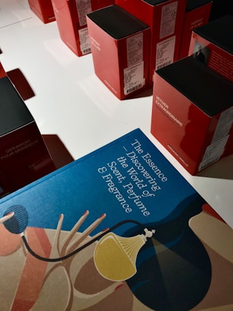 A collection of red and black boxes are neatly arranged on a white surface. In the foreground, there's a blue book titled 'The Essence Discovering the World of Scent, Perfume & Fragrance' with abstract art featuring a stylized perfume bottle and decorative motifs.