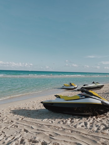 Jet skis are parked on a sandy beach with turquoise ocean water in the background. The sky is clear with a few scattered clouds.