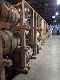 A storage area contains numerous wooden barrels arranged in rows on sturdy wooden supports. Soft lighting from exposed bulbs hangs above, casting a warm glow over the scene. The room has a spacious, industrial feel with its high ceilings and concrete floors.