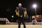 A person stands on an outdoor sports court at night, carrying two large duffle bags labeled 'Spikeball Pro.' The court is lit by bright overhead lights and features a basketball hoop and a fence.