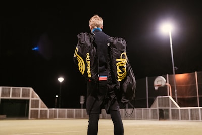 A person stands on an outdoor sports court at night, carrying two large duffle bags labeled 'Spikeball Pro.' The court is lit by bright overhead lights and features a basketball hoop and a fence.
