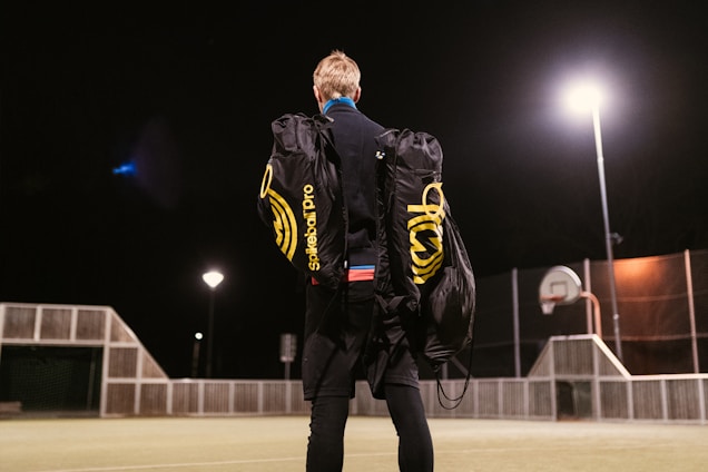 A person stands on an outdoor sports court at night, carrying two large duffle bags labeled 'Spikeball Pro.' The court is lit by bright overhead lights and features a basketball hoop and a fence.