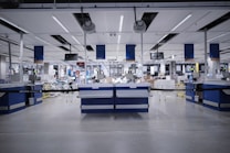 A spacious, modern warehouse or industrial interior with high ceilings and a sleek, organized layout. The space features several workstations equipped with monitors, barcode scanners, and storage areas. The setting is well-lit and appears clean and efficient, suggesting an environment designed for streamlined operations.