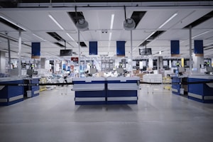 A spacious, modern warehouse or industrial interior with high ceilings and a sleek, organized layout. The space features several workstations equipped with monitors, barcode scanners, and storage areas. The setting is well-lit and appears clean and efficient, suggesting an environment designed for streamlined operations.