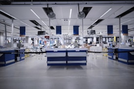 A spacious, modern warehouse or industrial interior with high ceilings and a sleek, organized layout. The space features several workstations equipped with monitors, barcode scanners, and storage areas. The setting is well-lit and appears clean and efficient, suggesting an environment designed for streamlined operations.