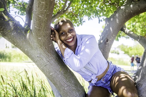A person is joyfully perched on a tree branch, surrounded by lush green leaves. They are wearing a light blue shirt and denim shorts, radiating happiness with a broad smile.
