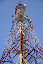 A tall metal telecommunications tower with a lattice structure dominates the landscape against a clear blue sky. Various antennas and receivers are mounted along its length for signal transmission.