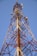 A tall metal telecommunications tower with a lattice structure dominates the landscape against a clear blue sky. Various antennas and receivers are mounted along its length for signal transmission.
