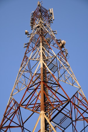 A tall metal telecommunications tower with a lattice structure dominates the landscape against a clear blue sky. Various antennas and receivers are mounted along its length for signal transmission.