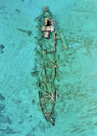 An aerial view of a sunken shipwreck surrounded by clear turquoise water. The ship's metal structure is partially visible beneath the water's surface, with rusted sections protruding above. The surrounding area is serene, with a few swimmers nearby exploring the site.