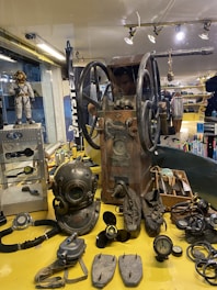 A collection of vintage diving equipment displayed on a yellow surface including a diving helmet, breathing apparatus, propeller, and gauges. An old machine with large wheels is positioned centrally. The background shows various nautical items and apparel.