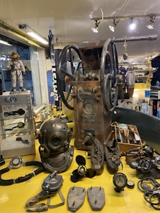 A collection of vintage diving equipment displayed on a yellow surface including a diving helmet, breathing apparatus, propeller, and gauges. An old machine with large wheels is positioned centrally. The background shows various nautical items and apparel.