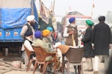 A group of men wearing turbans and traditional attire are gathered in an outdoor setting, engaged in conversation. Some are seated on plastic chairs while others stand around them. The backdrop features a large truck with a blue covering and other vehicles, amidst banners and flags.