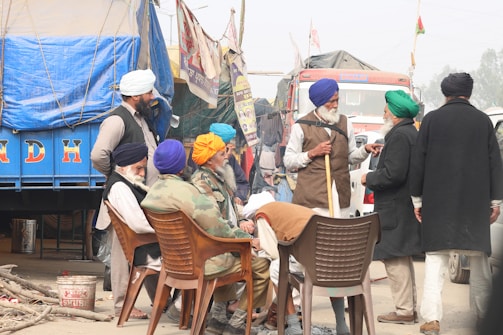 A group of men wearing turbans and traditional attire are gathered in an outdoor setting, engaged in conversation. Some are seated on plastic chairs while others stand around them. The backdrop features a large truck with a blue covering and other vehicles, amidst banners and flags.