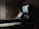 A dimly lit music studio featuring a keyboard in the foreground and a computer setup with dual monitors displaying audio software. Large speakers are positioned on either side of the monitors, and wooden acoustic panels cover the walls.