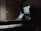 A dimly lit music studio featuring a keyboard in the foreground and a computer setup with dual monitors displaying audio software. Large speakers are positioned on either side of the monitors, and wooden acoustic panels cover the walls.