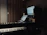 A dimly lit music studio featuring a keyboard in the foreground and a computer setup with dual monitors displaying audio software. Large speakers are positioned on either side of the monitors, and wooden acoustic panels cover the walls.