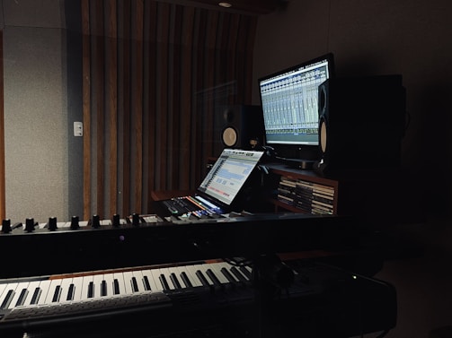 A dimly lit music studio featuring a keyboard in the foreground and a computer setup with dual monitors displaying audio software. Large speakers are positioned on either side of the monitors, and wooden acoustic panels cover the walls.