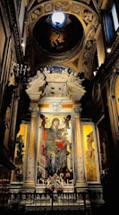 An ornate religious mural dominates the center, depicting a seated figure with a halo, likely a religious icon, surrounded by intricate architectural elements. The scene is framed by decorative columns and rich gilded patterns. A bouquet of flowers is placed at the base of the mural, and dim lighting enhances the sacred atmosphere.