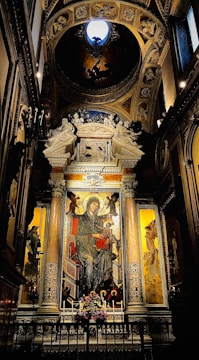 An ornate religious mural dominates the center, depicting a seated figure with a halo, likely a religious icon, surrounded by intricate architectural elements. The scene is framed by decorative columns and rich gilded patterns. A bouquet of flowers is placed at the base of the mural, and dim lighting enhances the sacred atmosphere.