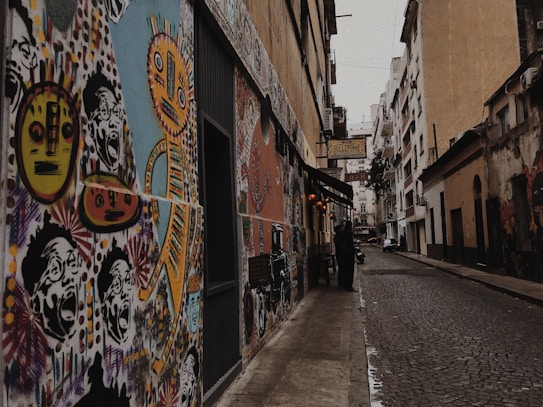 Vibrant street art covers the walls of a narrow alley, featuring colorful abstract faces and figures in a lively urban setting. The scene includes cobblestone pavement and buildings that line both sides of the alley. A sign hangs above a small business entrance.