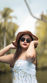 A person wearing a sunhat and sunglasses stands against a blurred background of greenery, including palm trees. They are dressed in a strapless white top with floral patterns, and their long hair falls over their shoulders. The image has an overall tropical and summery feel.