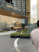 A modern glass building with an Apple logo on the front, flanked by high-rise structures. People are gathered outside, sitting on benches around a landscaped area. The scene is urban with a contemporary architectural design.