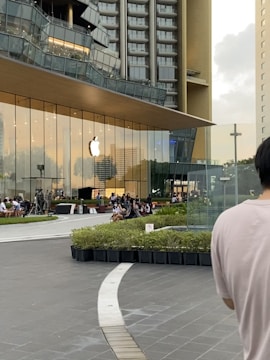 A modern glass building with an Apple logo on the front, flanked by high-rise structures. People are gathered outside, sitting on benches around a landscaped area. The scene is urban with a contemporary architectural design.