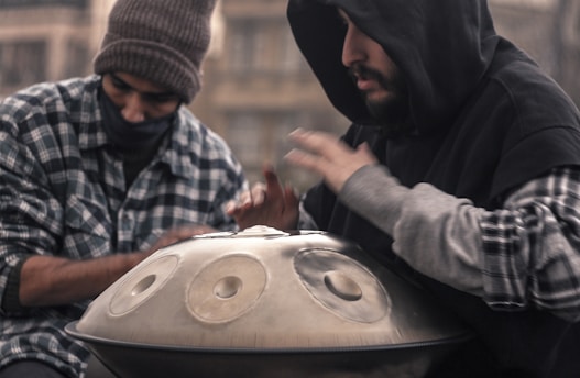 Two individuals, both wearing flannel shirts and hooded jackets, are engaged in playing a handpan. One of them is captured in motion, striking the instrument with their hands. The background is slightly blurred, giving the scene a sense of focus and intimacy on the musicians.