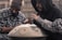 Two individuals, both wearing flannel shirts and hooded jackets, are engaged in playing a handpan. One of them is captured in motion, striking the instrument with their hands. The background is slightly blurred, giving the scene a sense of focus and intimacy on the musicians.