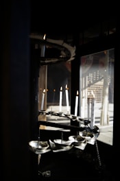 Several white candles are positioned in a spiral formation within a dark, enclosed space. They are lit, casting a warm glow, and there are reflections visible in a glass window nearby.