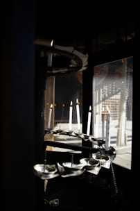 Several white candles are positioned in a spiral formation within a dark, enclosed space. They are lit, casting a warm glow, and there are reflections visible in a glass window nearby.