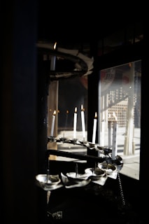 Several white candles are positioned in a spiral formation within a dark, enclosed space. They are lit, casting a warm glow, and there are reflections visible in a glass window nearby.